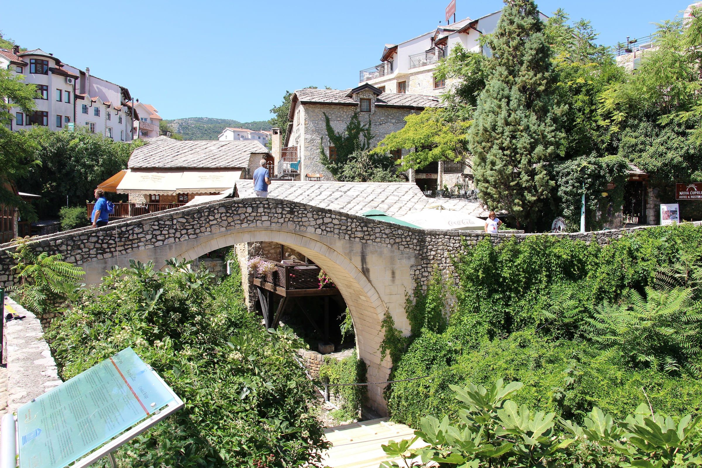 The small, steep stone arch of the Crooked Bridge over a stream, surrounded by greenery and old houses.