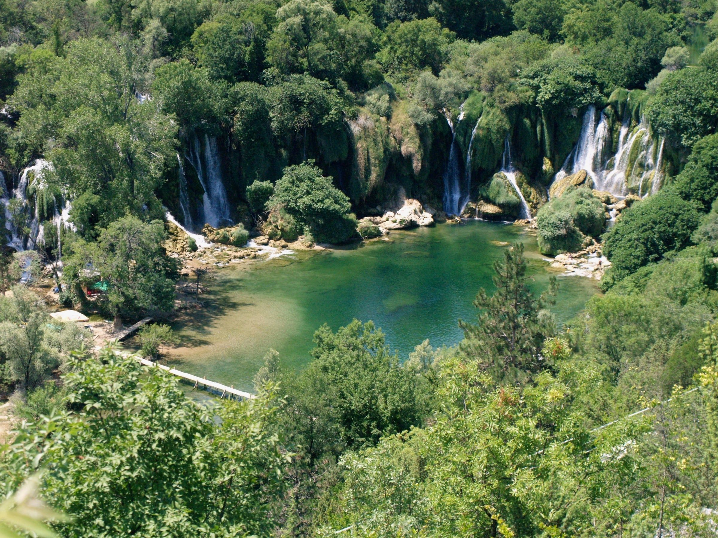 Kravice waterfalls seen from above, a semicircle of cascades falling into a green pool ringed by trees.