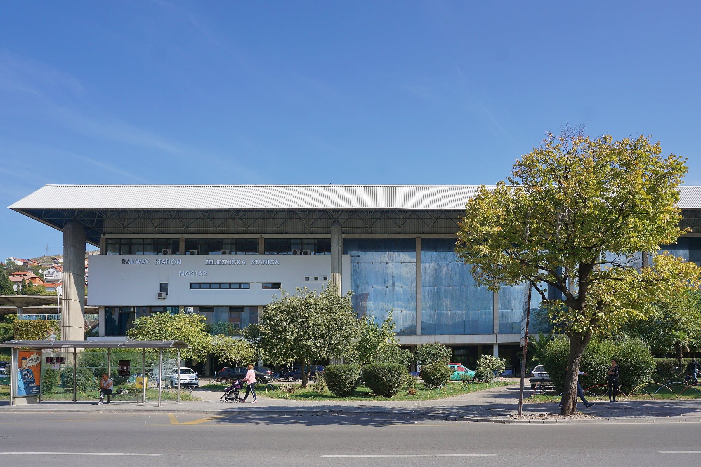The modernist Mostar railway station building, low and glass-fronted, with trees and a road in front.